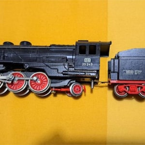 May include: A black model train with red wheels and accents, connected to a black tender car with red wheels. The train is set against a yellow background.