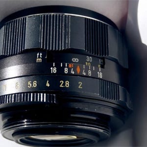 May include: A black camera lens with a silver ring and a depth of field scale marked in feet and meters. The aperture settings are 16, 11, 8, 5.6, 4, 2.8, and 2.