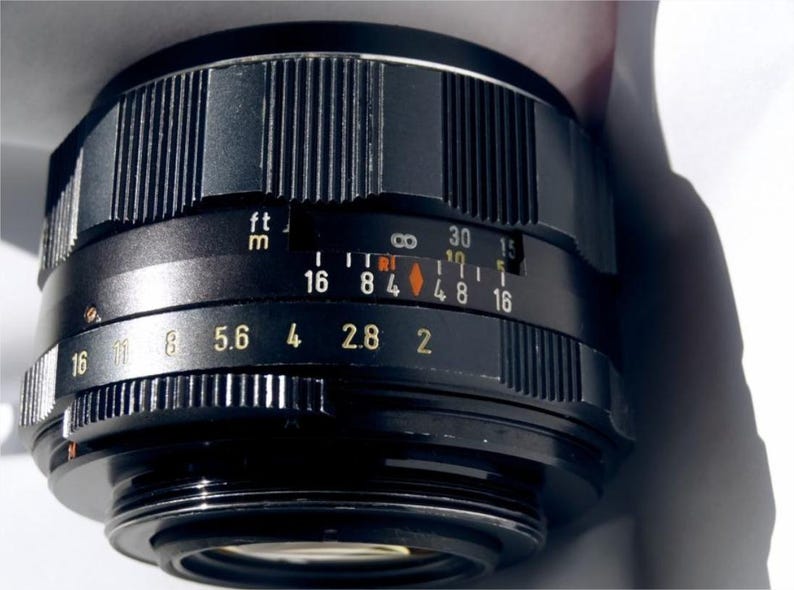 May include: A black camera lens with a silver ring and a depth of field scale marked in feet and meters. The aperture settings are 16, 11, 8, 5.6, 4, 2.8, and 2.