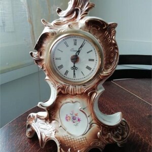 Puede incluir: Un reloj de chimenea de porcelana blanca con detalles dorados y un diseño floral. El reloj tiene una esfera redonda con números romanos negros y agujas negras.