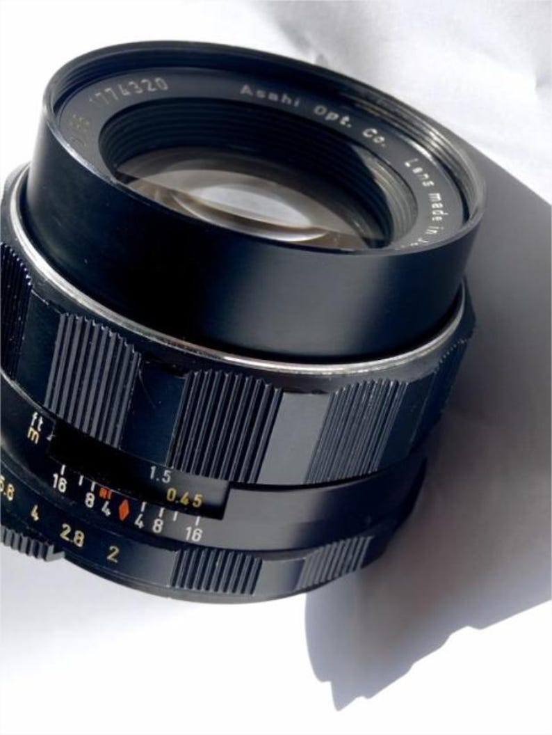 May include: A black camera lens with a silver ring and a black aperture ring. The aperture ring has markings for f/1.5, f/2, f/2.8, f/4, f/5.6, f/8, f/11, f/16, and f/0.45. The lens is labeled "Asahi Opt. Co. Lens Made in Japan".
