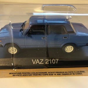 Puede incluir: Un coche modelo VAZ 2107 azul en una caja de plástico transparente. El modelo tiene ruedas blancas y una baca. El texto "VAZ 2107" es visible en la base. El embalaje incluye texto en rumano.