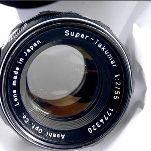 May include: A black camera lens with the text "Super-Takumar 1:2/55 1774320 Asahi Opt. Co. Lens made in Japan" printed on the side.