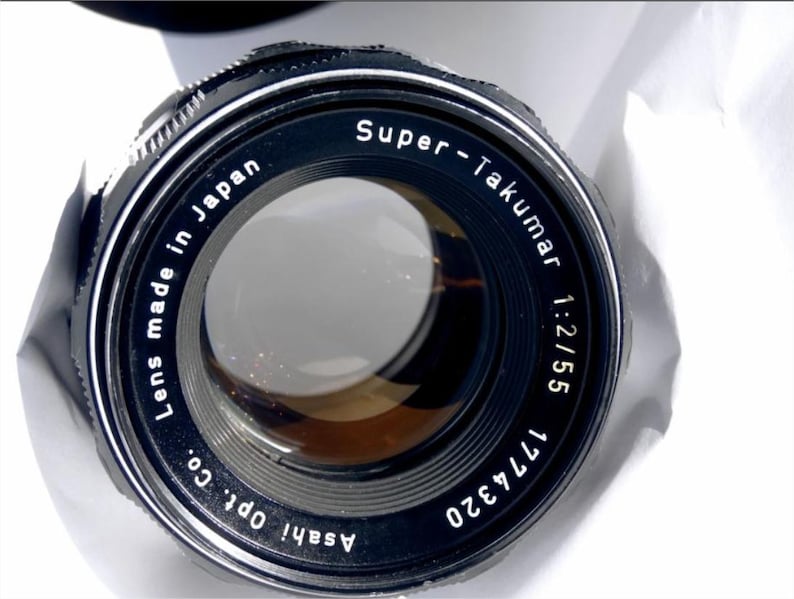 May include: A black camera lens with the text "Super-Takumar 1:2/55 1774320 Asahi Opt. Co. Lens made in Japan" printed on the side.