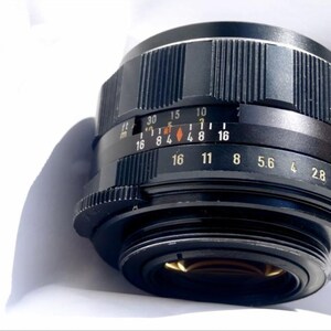 May include: A black camera lens with a silver aperture ring and depth of field markings. The lens has a black metal body and a silver ring with markings for aperture settings from f/2 to f/16.