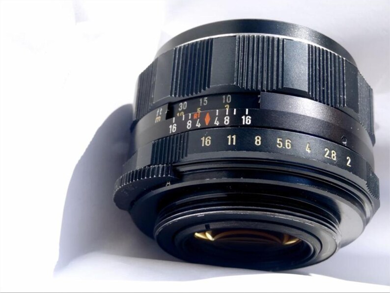 May include: A black camera lens with a silver aperture ring and depth of field markings. The lens has a black metal body and a silver ring with markings for aperture settings from f/2 to f/16.