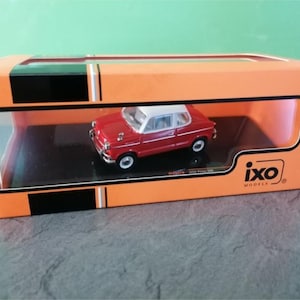 May include: A red and white model car in an orange display box. The miniature car has a white roof and small white wheels. The box has a clear plastic window and the "ixo MODELS" logo.