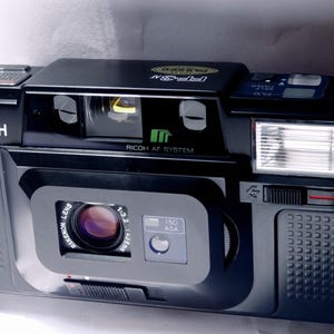 Peut inclure: Appareil photo argentique Ricoh AF-5M noir. L'appareil photo est équipé d'un objectif RIKENON avec une ouverture de 1:3.2 f=35mm. Il dispose d'un flash intégré, de réglages ISO/ASA et du système Ricoh AF. L'appareil est en bon état.