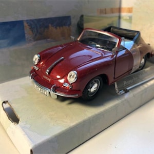 May include: A miniature red Porsche convertible car with a black interior and silver detailing. The model is presented in a box. The car features a number plate and the word "Porsche" is visible on the front.