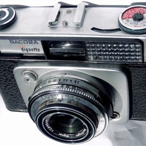 Puede incluir: Una cámara de película de 35 mm de color plateado y negro con una cubierta de imitación de cuero negro. La cámara tiene una lente plateada con un anillo negro alrededor. La lente tiene el texto "Reutlingen" y "Digimar" en ella. La cámara tiene una parte superior plateada con una perilla negra y un dial plateado con el texto "Color" en él.