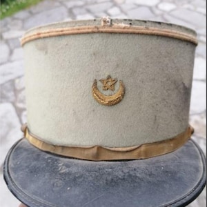 Puede incluir: Sombrero militar vintage verde claro con ala oscura. Presenta un emblema dorado de luna creciente y estrella. Una banda dorada rodea la base y una fina banda marrón claro adorna la parte superior.