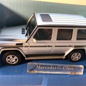 Könnte beinhalten: Ein silbernes Spielzeugautomodell eines Mercedes G-Klasse SUV. Das Auto hat einen schwarzen Innenraum und eine schwarze Plastikbasis mit dem Aufdruck "Mercedes G-Klasse".