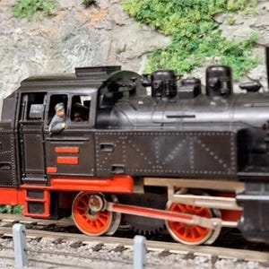 Peut inclure: Un modèle réduit de train noir avec des détails rouges, notamment sur les roues. Une figurine est visible dans la cabine du conducteur. Le train est posé sur une voie ferrée miniature, devant un décor de colline rocheuse.