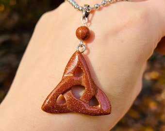 May include: A gold-toned chain necklace with a brown, sparkly, triquetra pendant. The pendant is made of a stone with a speckled texture.