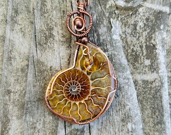 May include: A copper wire wrapped pendant featuring a fossilized ammonite with a golden brown color. The pendant is hanging from a copper chain on a wooden background.