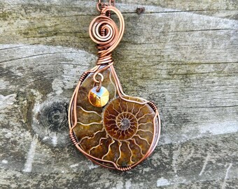 May include: A copper wire wrapped pendant featuring a fossilized ammonite with a golden iridescent accent. The pendant is hanging from a copper chain against a wood background.
