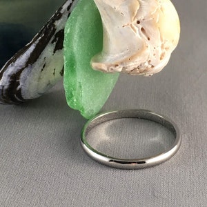 May include: A polished silver ring with a smooth, rounded band. The ring is displayed on a grey surface, with a green sea glass shard, a seashell, and a mussel shell in the background. The ring's simple design makes it suitable for everyday wear.