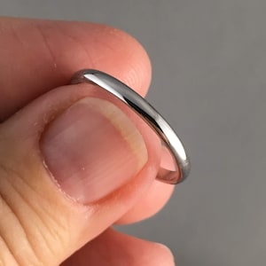 May include: A shiny, silver ring with a rounded band. The ring is held between two fingers against a grey background. The ring's simple design suggests it could be a wedding band or a minimalist piece of jewellery. The ring's band is approximately 2 mm wide.