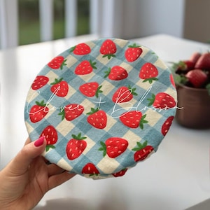 May include: A round, fabric food cover featuring a blue and white gingham pattern with red strawberries. The cover has the text "Love + Blossom" in a cursive font. The cover is held in a person's hand.