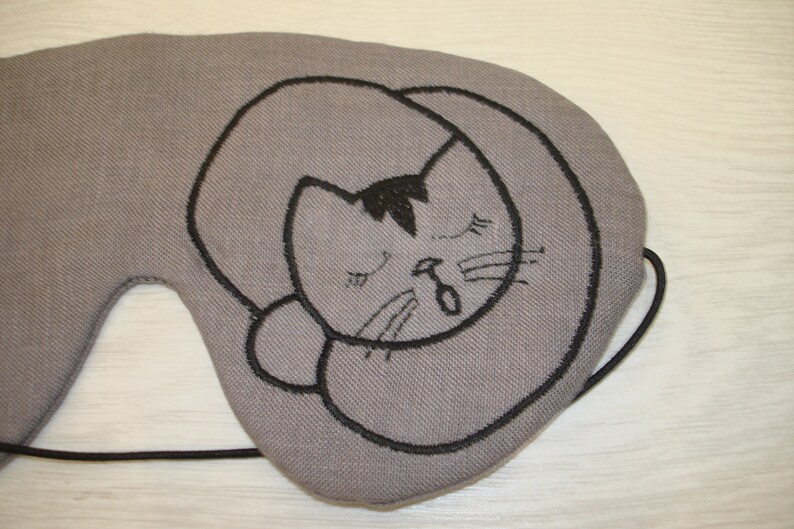 May include: Gray eye mask with a black embroidered sleeping cat design.