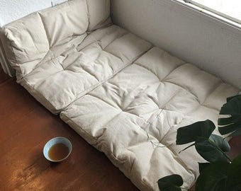 Puede incluir: Un juego de cojines de suelo beige y con mechones dispuestos en forma de L, cerca de una ventana. Los cojines están sobre un suelo de madera. Un pequeño cuenco con líquido está en el suelo. Una planta verde está en la esquina.