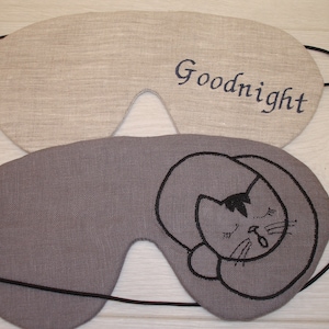May include: Two sleep masks made of linen fabric. One is beige with the word "Goodnight" embroidered in blue thread. The other is gray with a black embroidered image of a cat sleeping.