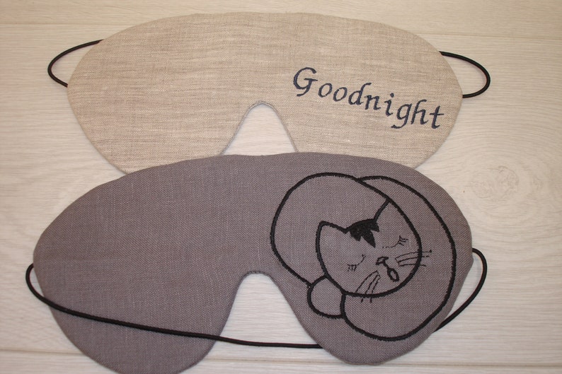 May include: Two sleep masks made of linen fabric. One is beige with the word "Goodnight" embroidered in blue thread. The other is gray with a black embroidered image of a cat sleeping.