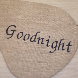May include: A beige linen sleep mask with an elastic strap. The mask has the word "Goodnight" embroidered in black thread.