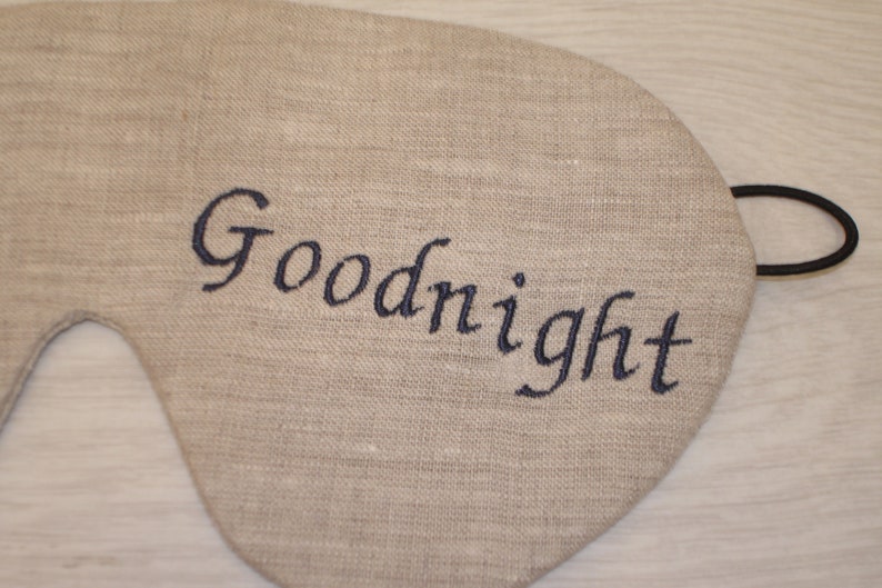 May include: A beige linen sleep mask with an elastic strap. The mask has the word "Goodnight" embroidered in black thread.