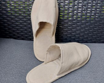Può includere: Due pantofole beige a punta aperta. Le pantofole sono realizzate in un tessuto morbido e strutturato. Una pantofola è in posizione verticale e l'altra è sdraiata. Lo sfondo è una superficie grigio scuro e un motivo intrecciato nero.