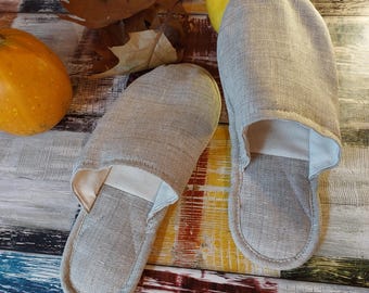 Può includere: Un paio di pantofole di lino grigie con fodera bianca. Le pantofole sono su una superficie di legno con uno sfondo colorato.