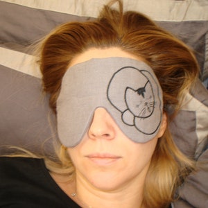 May include: A gray sleep mask with a black embroidered cat design. The mask is worn by a person with long, light brown hair.