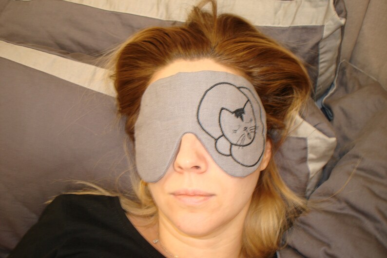 May include: A gray sleep mask with a black embroidered cat design. The mask is worn by a person with long, light brown hair.