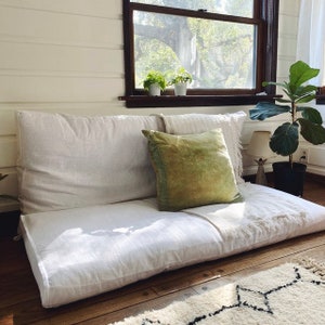 Op de afbeelding: Een vloerkussen met een grote witte linnen basis en rugleuning, aangevuld met een groen fluwelen kussen en een crèmekleurige deken. De scène speelt zich af bij een raam met planten, waardoor een gezellige, uitnodigende ruimte ontstaat.