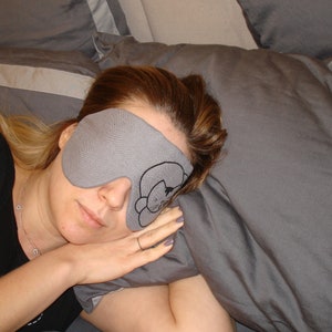 May include: A woman wearing a gray sleep mask with a cat design on a gray pillow. The mask is covering her eyes and she is sleeping.