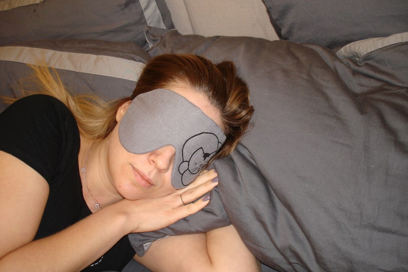 May include: A woman wearing a gray sleep mask with a cat design on a gray pillow. The mask is covering her eyes and she is sleeping.