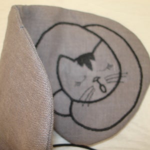 May include: A gray eye mask with a black embroidered design of a sleeping cat. The mask has a strap to go around the head.