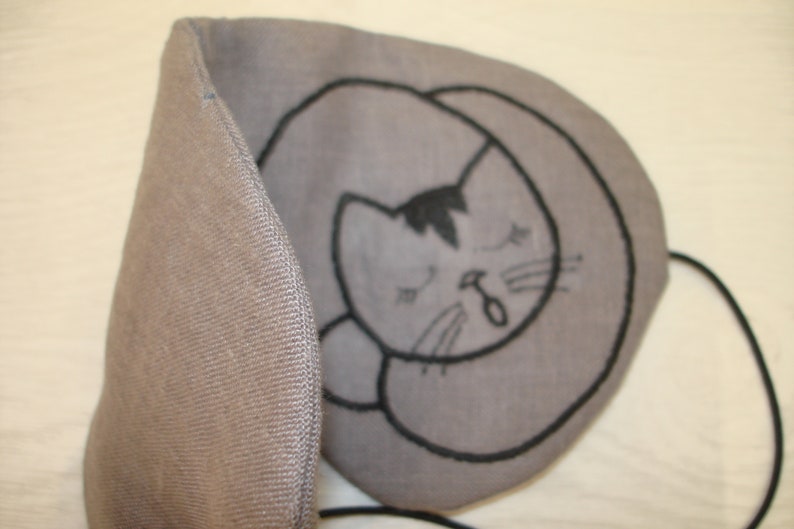 May include: A gray eye mask with a black embroidered design of a sleeping cat. The mask has a strap to go around the head.