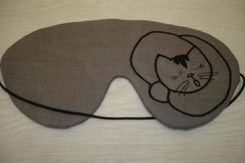 May include: Gray linen sleep mask with a black embroidered sleeping cat design.