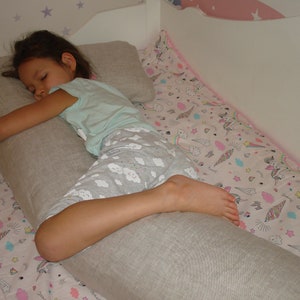 Puede incluir: Un niño dormido en una cama, usando una almohada larga y gris para el cuerpo y una almohada más pequeña. La cama tiene una funda nórdica estampada con unicornios y arcoíris. La pared es blanca con pegatinas de estrellas.