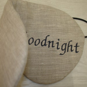May include: A beige linen sleep mask with an elastic band and the word "goodnight" embroidered in black thread.