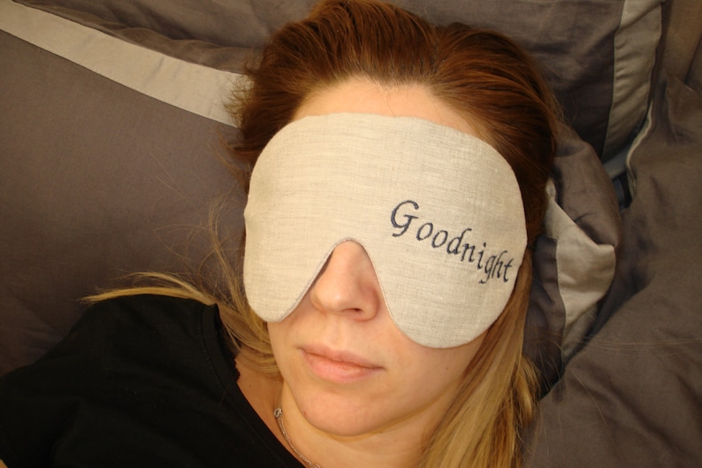 May include: A close-up of a person wearing a light brown sleep mask with the word "Goodnight" embroidered on it in dark blue thread. The person is lying down on a gray pillow.