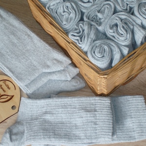 Puede incluir: Calcetines de color gris claro, algunos enrollados en una cesta de mimbre, otros desplegados. También hay una etiqueta de madera con el texto "EcoStyle Organic Buying" y un logotipo de hoja. Los calcetines son de tejido acanalado. La imagen sugiere productos orgánicos.
