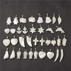 Może przedstawiać: Kolekcja wisiorków z masy perłowej w różnych kształtach, w tym serca, krzyże, ryby i stopa. Każdy wisiorek ma srebrne kółeczko, a niektóre zdobione są małymi, przezroczystymi kryształkami. Wisiorki są prezentowane na ciemnoszarej powierzchni.