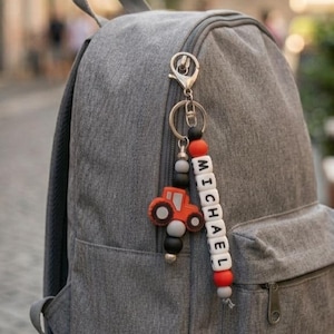 Könnte beinhalten: Ein grauer Rucksack mit einem roten und schwarzen Traktor-Schlüsselanhänger. Der Schlüsselanhänger trägt den Namen "MICHAEL" in weißen Buchstabenperlen. Der Rucksack steht auf einem Holztisch.