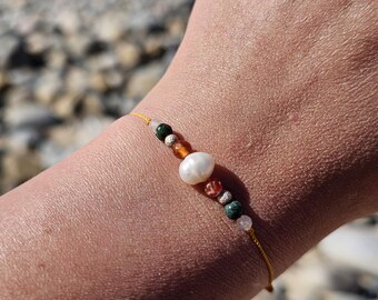 May include: A delicate bracelet featuring a string of colorful beads and a large, white pearl. The beads include green, orange, and clear stones. The bracelet is on a yellow string.