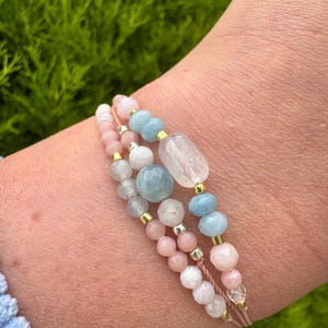 May include: Three-strand bracelet featuring pink, white, and light blue gemstone beads. The bracelet includes a variety of bead shapes and sizes, with gold-colored metal accents. The gemstones are strung on a light pink cord.