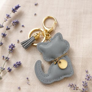 Puede incluir: Un llavero con forma de gato de color gris hecho de material liso con costuras visibles en los bordes. Cuenta con un cierre de metal dorado, una borla gris a juego y un pequeño dije circular dorado unido por una delicada cadena dorada.