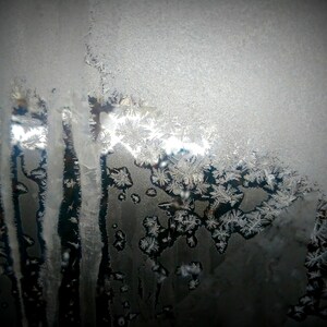 Puede incluir: Un primer plano de un panel de ventana cubierto de intrincados patrones de escarcha. La escarcha es blanca y delicada, creando un diseño hermoso y abstracto. La luz brilla a través de la escarcha, creando un brillo suave y etéreo.