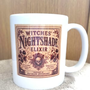 May include: A white ceramic mug featuring a vintage-style rectangular label with the text Witches' Nightshade Elixir, 1913, and Prepared by Old Salem Apothecary. The design includes a central skull illustration, botanical motifs, and warning text.